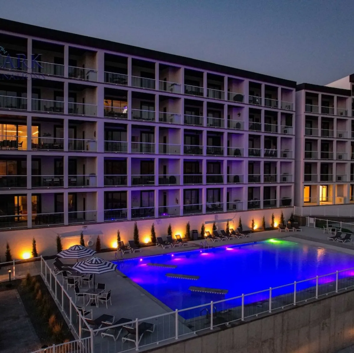 An exterior nighttime shot of an illuminated swimming pool with lounge chairs and visible balconies above. The luxurious setting invites swimming, socializing, or relaxing by the pool under the stars, with a sense of resort-style leisure.