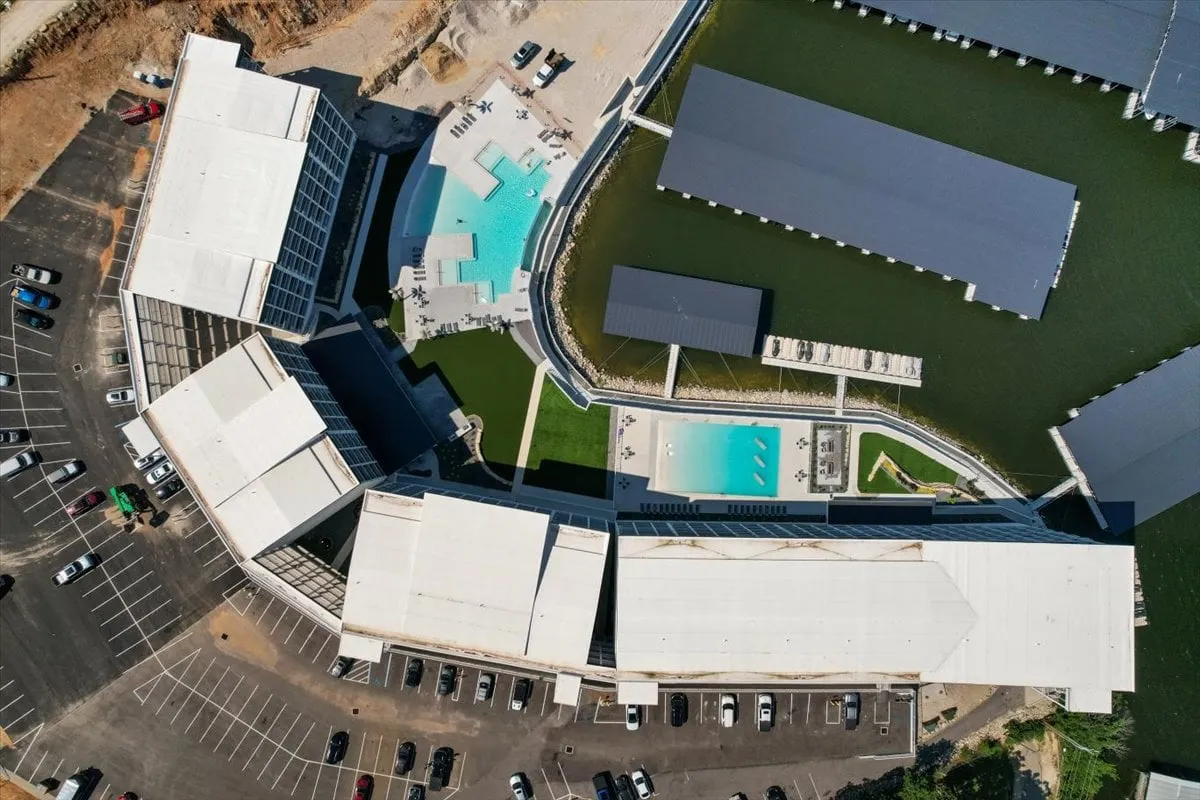 A vibrant exterior image featuring a swimming pool, modern lakefront architecture, and surrounding water views. The scene is lively, with sunbathing spots and hints of boating or water activities in the background.