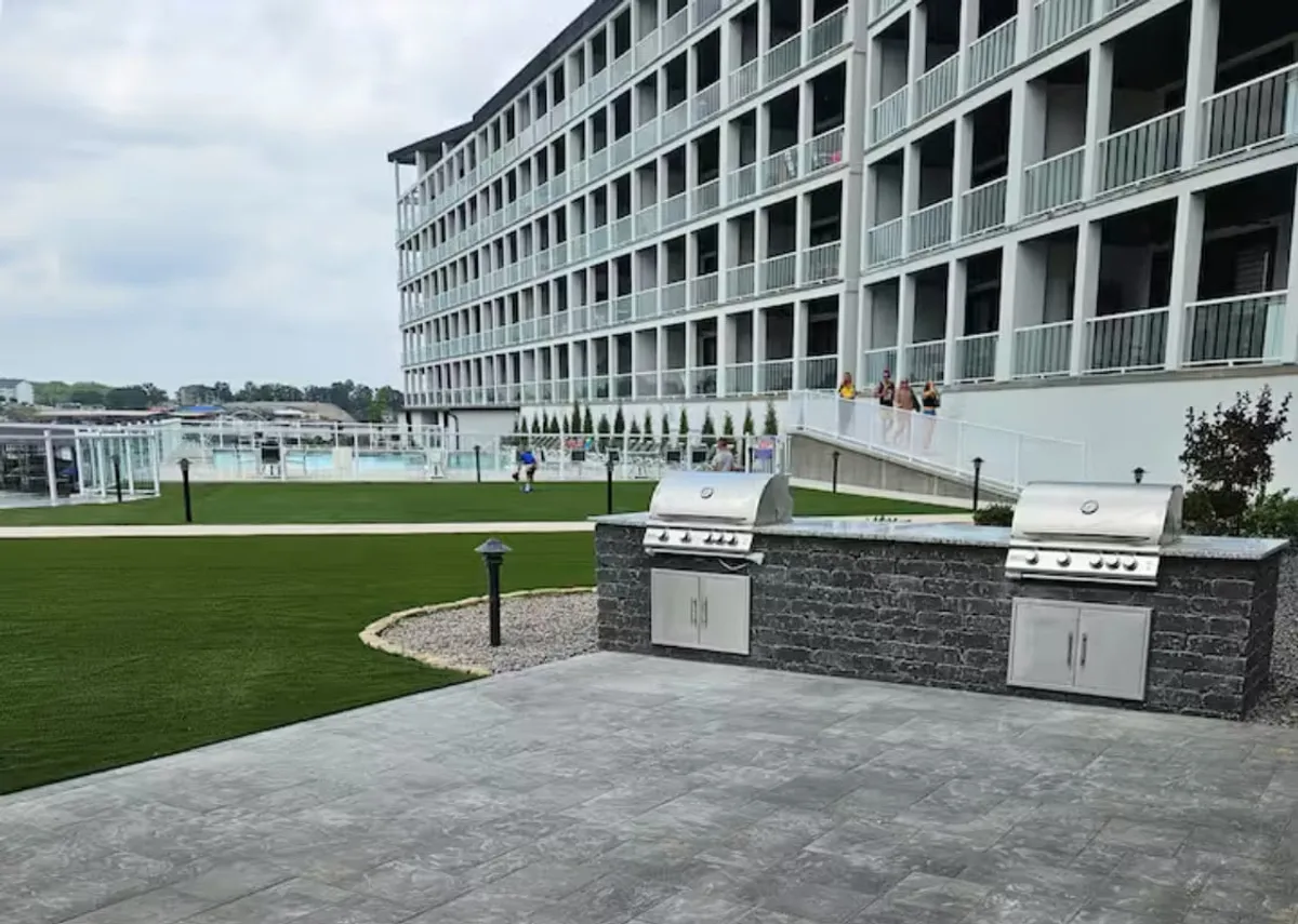 A cheerful outdoor grilling area with a well-maintained lawn, visible balconies, and glimpses of the pool in the background. The scene suggests communal gatherings and family celebrations in a sunny, open-air environment.