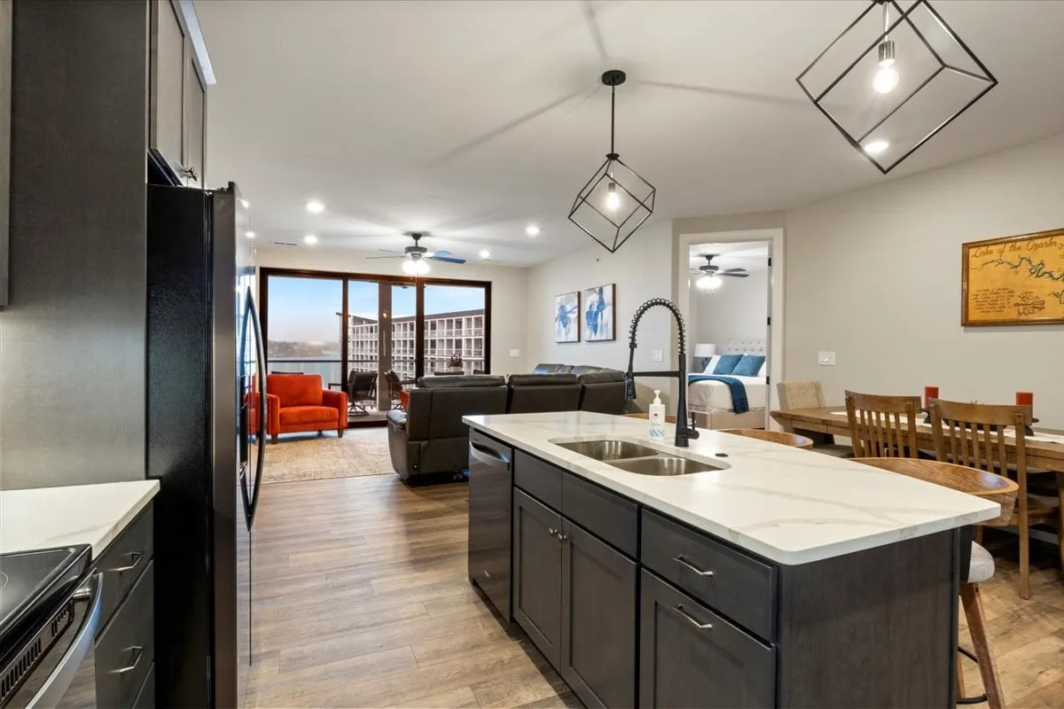 A wide-angle view of the open-plan living area, showing the kitchen island, dining table, and living area with plush seating. Large windows fill the space with natural light and offer scenic views of the lake, while sleek cabinetry and modern appliances set a contemporary tone. The flow between spaces is seamless, ideal for group activities.