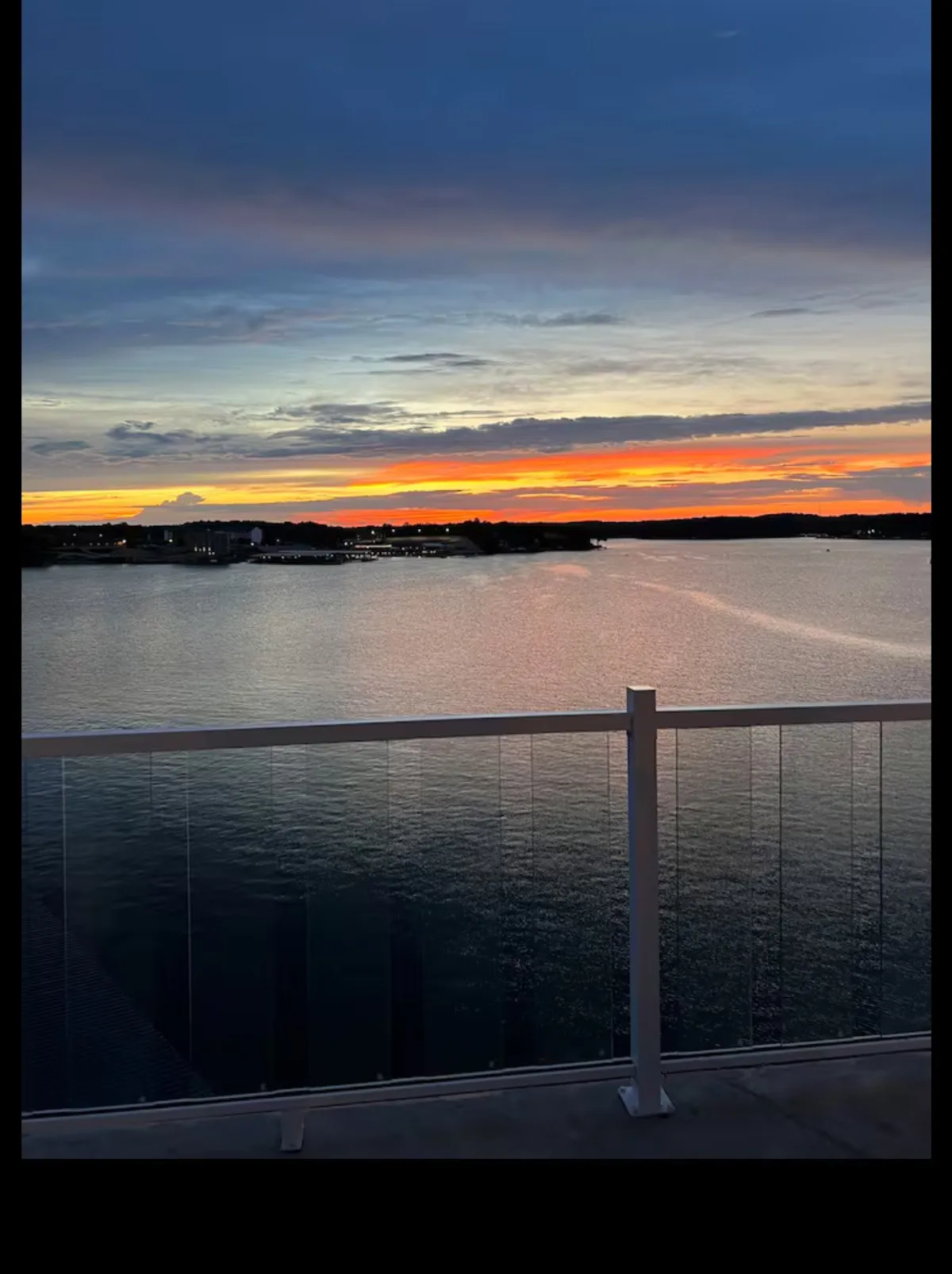 A stunning view of the sunset sky over calm lake waters, with a distant shoreline visible. The vantage point suggests the perspective from a balcony or lakeside spot, evoking tranquility and connection to nature.