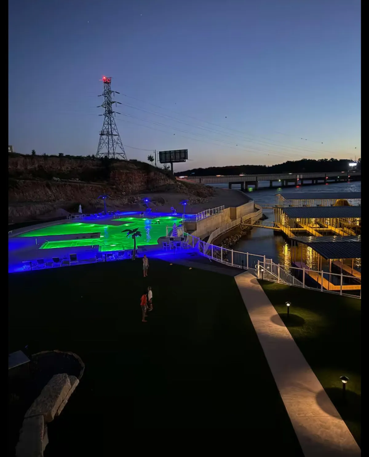 An illuminated swimming pool at dusk, with people socializing and enjoying the inviting outdoor space. Decorative lighting and reflections on the water create a festive, lively ambiance, hinting at evening celebrations and group gatherings.
