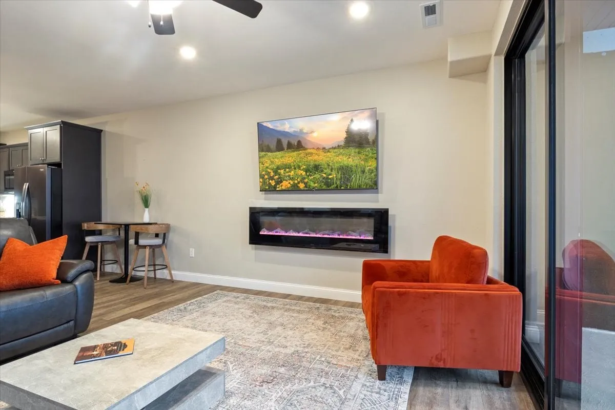 A cozy living area with an inviting orange accent chair, a modern electric fireplace, large wall-mounted TV, and a decorative table. The soft ambiance and stylish touches create a comfortable spot for socializing, relaxing, or sharing stories by the fire.