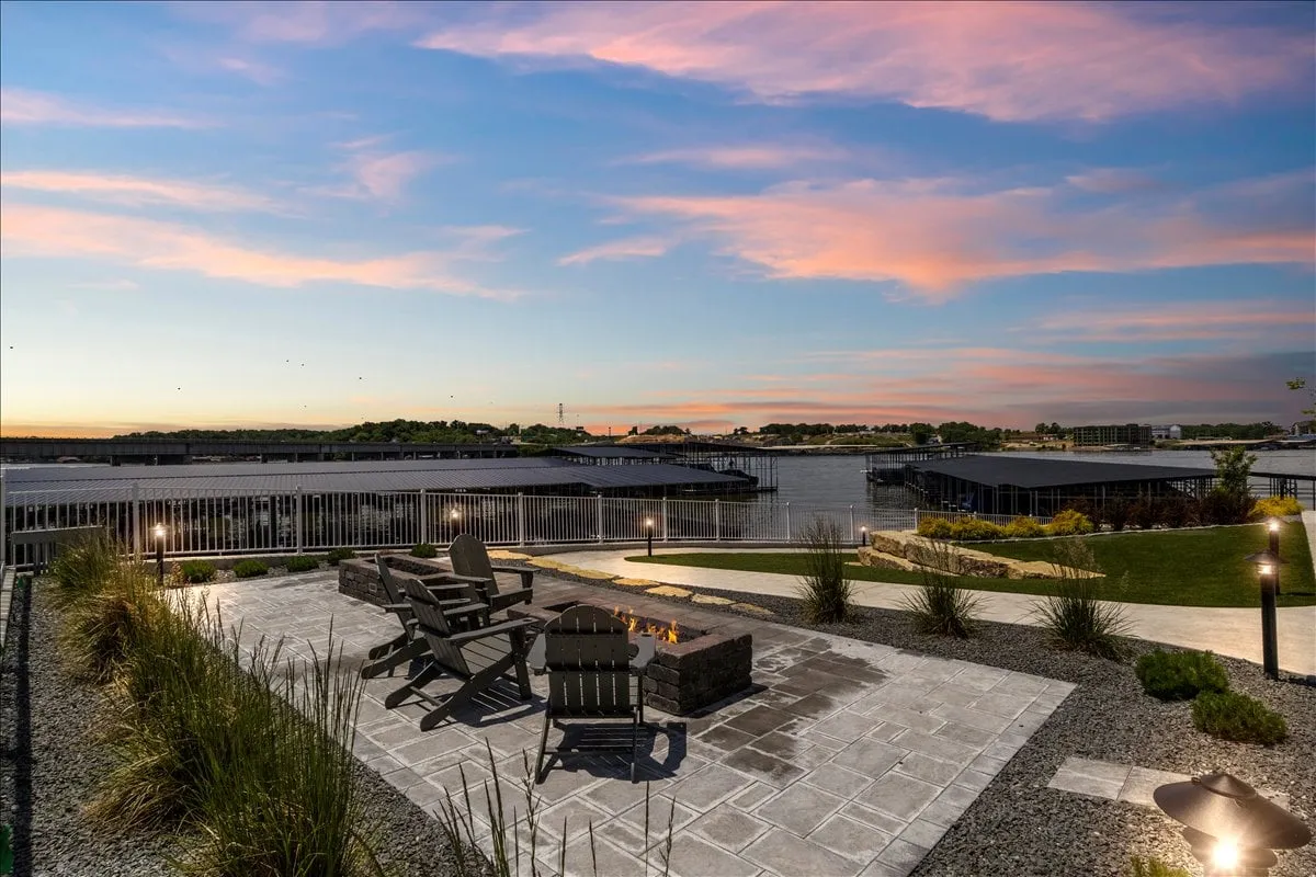 A cozy outdoor fire pit area with Adirondack chairs facing a tranquil waterfront. The landscaped garden and golden sunset create a warm, inviting atmosphere ideal for evening gatherings and stargazing.