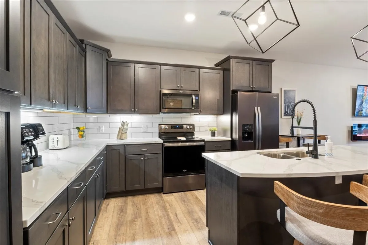 A contemporary kitchen featuring a spacious island, sleek dark cabinetry, stainless steel appliances, and geometric light fixtures. The countertops are uncluttered, suggesting the kitchen is fully stocked and ready for guests, embodying convenience and modern comfort.