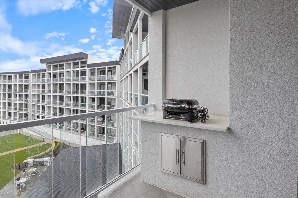 A view of a balcony showcasing a grill on the countertop, balcony railing, and adjacent buildings. The scene suggests a space ideal for barbecuing or enjoying a meal outdoors, with hints of casual leisure and sunset ambiance.